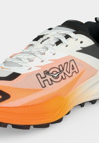 Κοντινό πλάνο ενός παπουτσιού τρεξίματος Hoka με λευκά κορδόνια, πορτοκαλί σταδιακή σόλα και μαύρες και λευκές λεπτομέρειες στο επάνω μέρος.