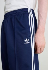 Námořnické modré sportovní kalhoty s elastickým pasem, se třemi bílými bočními pruhy a bílým logem Adidas na levém stehně.