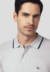 Jasnoszara koszulka polo z teksturowanej tkaniny, granatowym wykończeniem kołnierzyka oraz dwoma guzikami przy zapięciu. Posiada małe haftowane logo na klatce piersiowej.