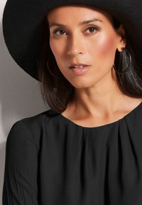 Schwarzes gefaltetes Oberteil mit rundem Ausschnitt, kombiniert mit einem breitkrempigen schwarzen Filzhut und großen schwarzen Creolen. Sanfte Textur.