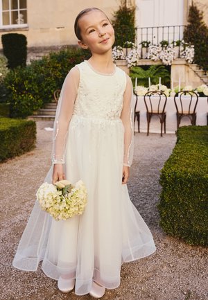 Junges Mädchen in langem weißen Kleid hält einen Strauß weißer Blumen und steht draußen nahe einem dekorierten Tisch und grünen Hecken.