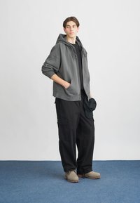 Felpa grigia con una vestibilità comoda, t-shirt nera sotto, pantaloni cargo neri e scarpe slip-on beige, modello in piedi su uno sfondo blu.