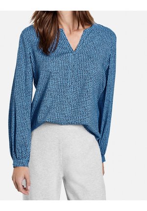 Femme portant un chemisier bleu à manches longues avec un motif à pois noirs et un pantalon gris clair, debout devant un fond blanc.