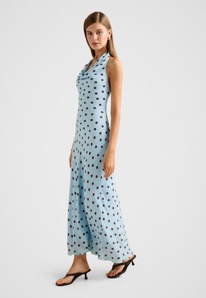 Femme portant une robe maxi bleu clair sans manches à pois noirs et des sandales à talons noires, debout devant un fond uni.