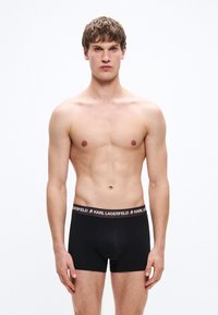 Μαύρα μποξέρ με εφαρμοστή σχεδίαση και ελαστική ζώνη μέσης με λογότυπο. Το μοντέλο είναι γυμνόστηθο, όρθιο με φόντο μονόχρωμο.