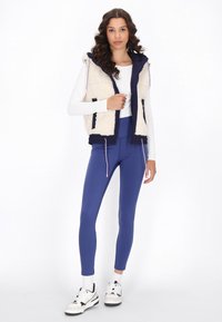 Gilet in pelliccia sintetica color crema con dettagli blu navy, abbinato a leggings blu reale e sneakers bianche. Il gilet presenta una chiusura con zip e coulisse.