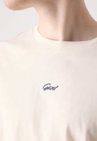Lys beige bomull t-skjorte med rund hals og korte ermer; har et lite, brodert mørkt logo på brystet.