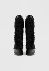 Črne krzneni škornji imajo dlakav teksturo in gladke črne gumijaste podplate, z visokim, strukturiranim zgornjim delom in zaobljenim dizajnom prstov.