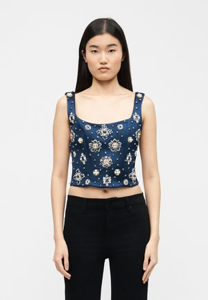 Femme portant un crop top sans manches bleu marine orné de grandes et petites décorations argentées et nacrées, associé à un pantalon noir.