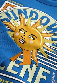 Niebieska tkanina ze stylizowanym słońcem z ludzką twarzą, otoczona tekstem zawierającym słowa "Mykonos" i "Sundown Venice" napisanymi pogrubioną czcionką.