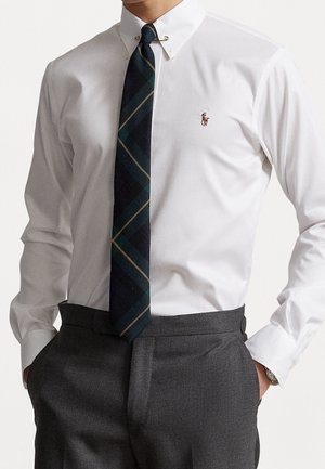 Mężczyzna w białej koszuli z haftowanym logo, ciemnym krawacie w kratę przypiętym agrafką oraz szarych spodniach garniturowych, z rękami w kieszeniach.