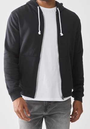 Homme portant un sweat à capuche noir zippé avec des cordons blancs sur un t-shirt blanc uni et un jean gris, debout devant un fond uni.