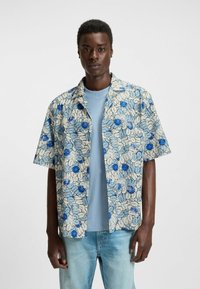 Lyhythihainen kukkapaidan, jossa on vaalea tausta sinisillä ja tummansinisillä kukilla, päällä vaaleansininen t-paita, yhdistetty vaaleisiin farkkuihin.