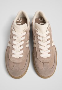Paire de baskets en daim beige avec lacets blancs et accents en cuir crème, vues de dessus sur un fond gris clair.