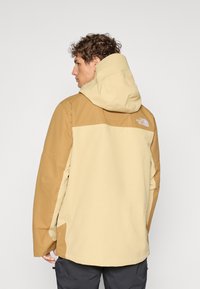Beež ja pruun välistingimustes jope kapuutsiga, tekstuurse kangaga, küljetaskute ja North Face logo'ga selja ülaosas.
