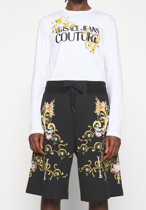 Osoba nosząca białą bluzkę z długim rękawem z napisem „Versace Jeans Couture” i żółtym kwiatowym wzorem, w połączeniu z czarnymi szortami ozdobionymi kolorowymi wzorami kwiatowymi.