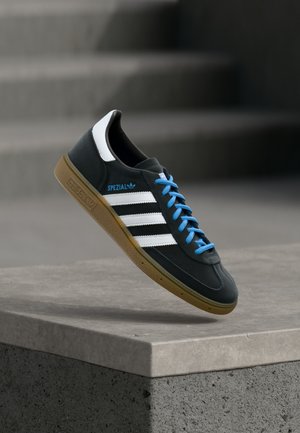 Μαύρο αθλητικό παπούτσι Adidas με λευκές ρίγες, μπλε κορδόνια και καουτσούκ σόλα να αιωρείται πάνω από γκρίζο τσιμεντένιο σκαλοπάτι σε απαλό φως.