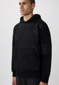 Sudadera negra hecha de tejido texturizado y suave, con un bolsillo frontal, paneles de malla en los hombros y un logo hexagonal en el pecho.