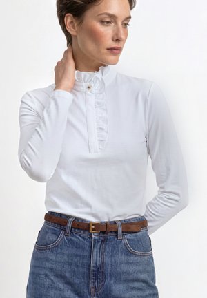 Woman wearing a white long-sleeve blouse with a ruffled high collar and blue jeans with a brown belt, hand touching her neck.