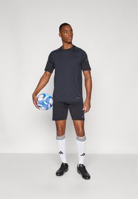 Zwart sportshirt met subtiele strepen, zwarte shorts, witte sokken met zwarte strepen en zwarte voetbalschoenen. Houdt een kleurrijke voetbals in de hand.