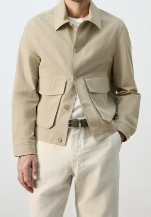 Mand iført beige jakke med knapper og to store forlommer, hvide bukser og hvid skjorte, står med den ene hånd i lommen.