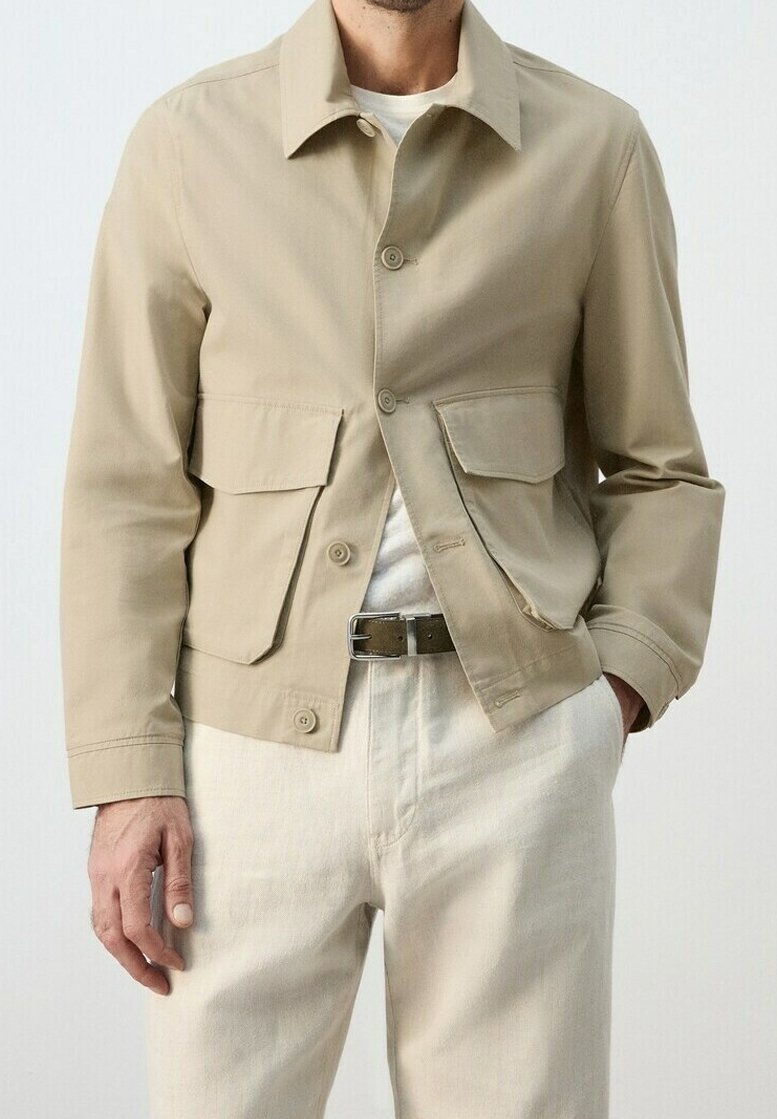 Homme portant une veste beige boutonnée avec deux grandes poches devant, pantalon blanc et chemise blanche, debout avec une main dans la poche.