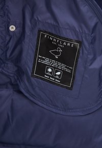 Etiqueta en chaqueta azul marino con la marca "Finnflare Est. 1965", icono de pato y texto que describe la calidez y flexibilidad del material de plumón de pato auténtico.
