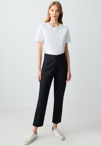 T-shirt blanc à col rond associé à un pantalon noir ajusté à taille haute. Le modèle porte des baskets blanches. Design simple et texture de tissu lisse.