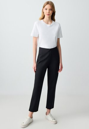 T-shirt bianco a girocollo abbinato a pantaloni neri a sigaretta a vita alta. Il modello indossa sneakers bianche. Design semplice e texture del tessuto liscia.
