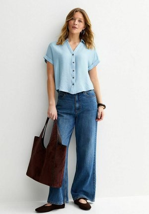 Młoda kobieta w jasnoniebieskiej koszuli z krótkim rękawem z zapięciem na guziki, szerokie niebieskie dżinsy, trzymająca dużą brązową torbę na ramię, stojąca na tle białej ściany.