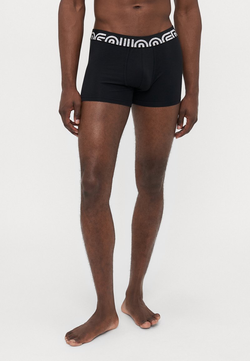Mann steht barfuß und trägt schwarze Boxershorts mit gemustertem Bund vor einem schlichten weißen Hintergrund.