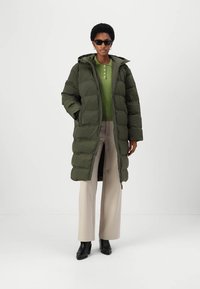 Green puffer coat with a hood, featuring a quilted texture and a straight cut. Worn over a light green top and cream-colored trousers.