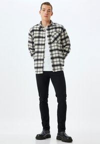 Camisa de franela a cuadros en blanco y negro con un bolsillo en el pecho, llevada sobre una camiseta blanca, combinada con pantalones ajustados negros y botas negras.