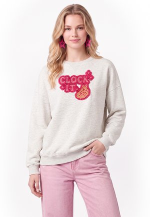 Ung kvinde med blondt hår iført en hvid sweatshirt med rød tekst ""Clock It"" og flammegrafik, lyserøde stjerneøreringe og lyserøde bukser.