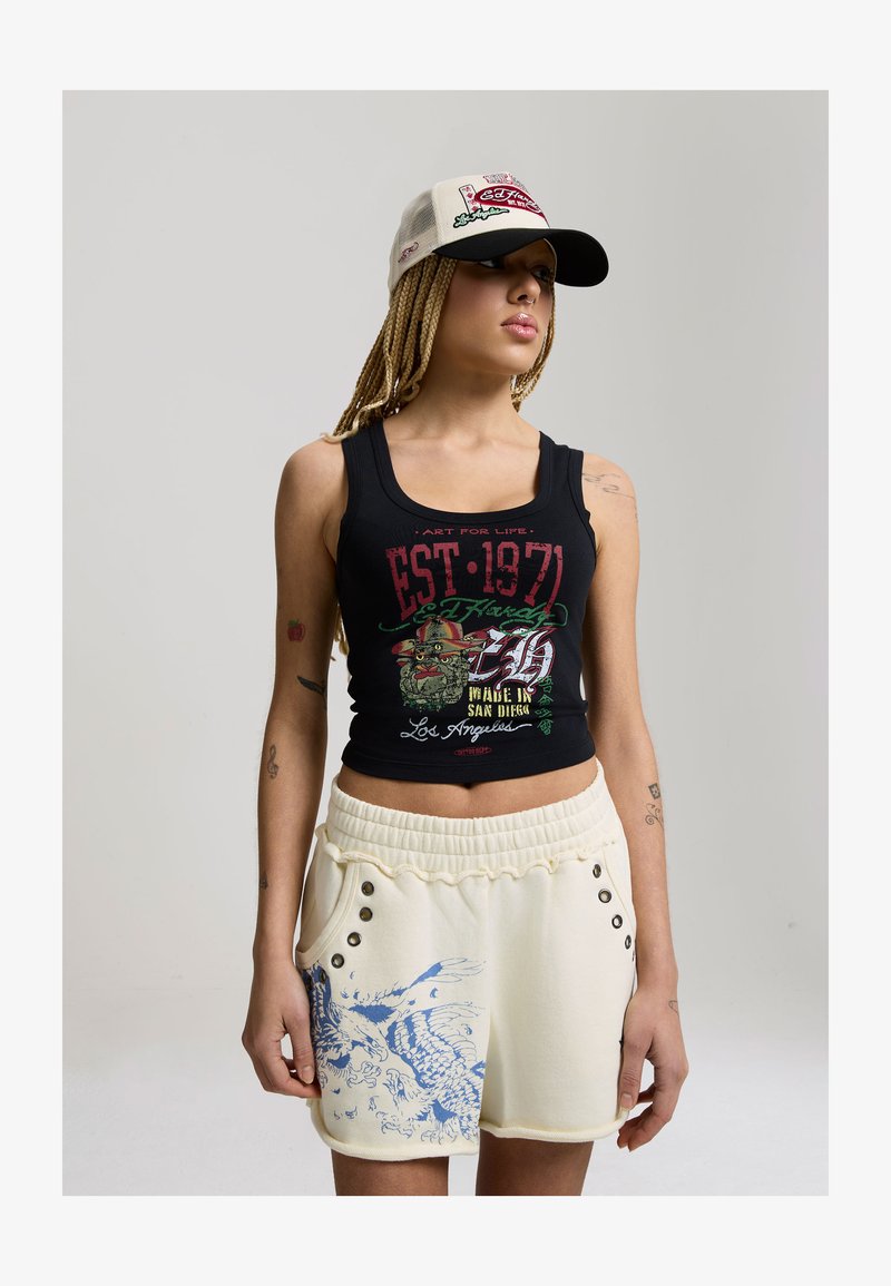 Frau mit geflochtenem Haar trägt ein schwarzes Tanktop mit Grafik, cremefarbene Shorts mit blauem Muster und eine beigem-schwarze Kappe, steht vor einem schlichten Hintergrund.