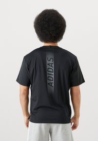 Must beebi lühikeste varrukatega T-särk, millel on vertikaalne hall "ADIDAS" logo ja graafikmuster seljal. Valmistatud siledast kangast, lihtne disain.