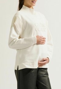 Crème-kleurige sweatshirt met een hoge ribbelkraag, gestructureerde schouderpanelen en een reliëflogo, gecombineerd met zwarte leren broek.