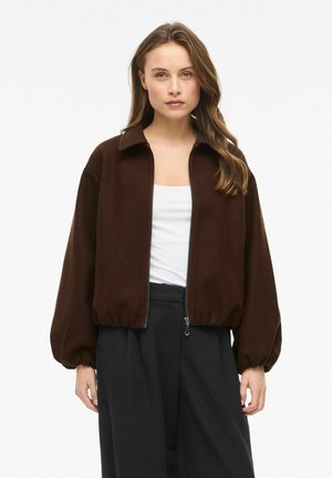 Femme portant une veste marron foncé zippée avec des manches bouffantes, un haut blanc et un pantalon noir taille haute, debout devant un fond blanc uni.