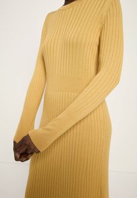 Frau, die ein senfgelbes, geripptes Strickkleid mit langen Ärmeln und taillierter Passform trägt, die Hände vorne zusammengelegt.