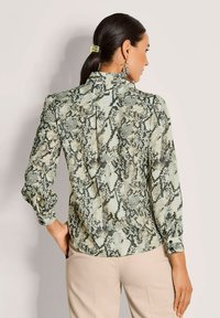 Frau mit dunklem Haar im Pferdeschwanz, die eine Bluse mit Schlangendruck und beige Hose trägt, von hinten vor einem schlichten Hintergrund gezeigt.