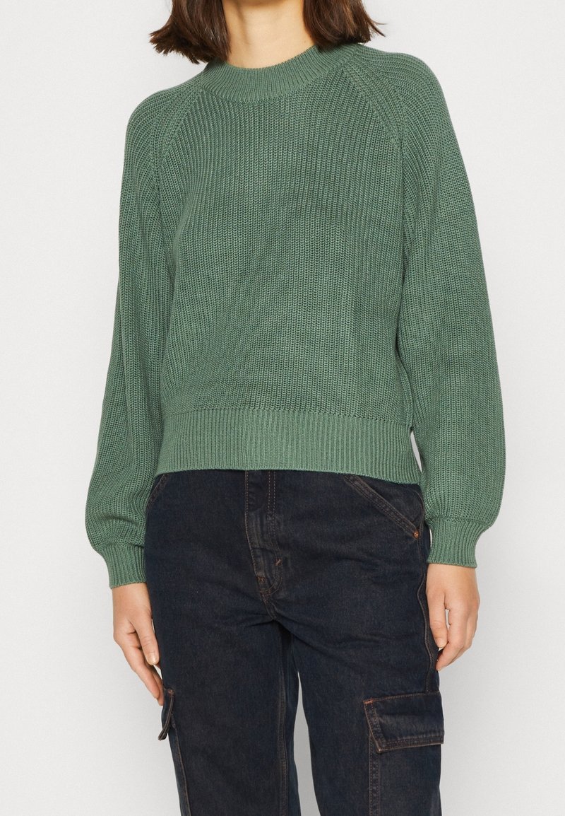 Personne portant un pull en maille vert avec poignets et ourlet côtelés, associé à un jean cargo bleu foncé avec poches latérales.