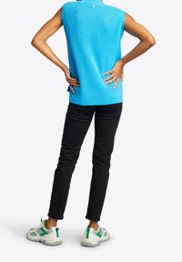 Blau gerippter ärmelloser Oberteil, schwarze slim-fit Jeans und weiße Sneakers mit grünen Akzenten. Das Modell steht mit Händen in den Hüften, von hinten gesehen.