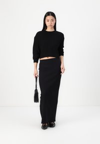 Μαύρος κοντός πουλόβερ με υφή πλέξης, συνδυασμένο με μακριά μαύρη φούστα. Τα αξεσουάρ περιλαμβάνουν μια μικρή μαύρη τσάντα και μαύρα παπούτσια.