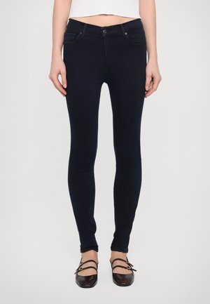 Dark blue skinny jeans with a smooth texture, mid-rise waist, front pockets, and a button closure. Worn with black flat shoes.