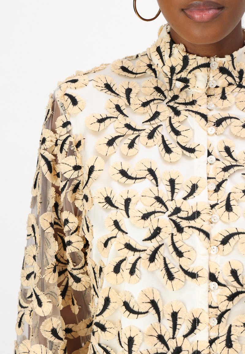 Bež cvjetni vez na prozirnoj tkanini s crnim naglascima. Ima visok ovratnik i gumbe sprijeda. Složen zamišljen uzorak.