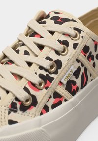 Μπέζ πάνινο αθλητικό παπούτσι με μαύρη και κόκκινη λεοπάρ εκτύπωση, μπεζ κορδόνια, χρυσά ρόδακες και μια μικρή λευκή υφασμάτινη ετικέτα «ONLY» στο πλάι.