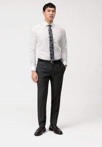 Chemise blanche, cravate noire à motifs, pantalon gris foncé, ceinture noire et mocassins foncés. Tissu lisse avec une coupe ajustée.