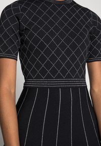 Robe en maille noire avec manches courtes, motif en losanges avec accents blancs, corsage ajusté et jupe à rayures verticales.