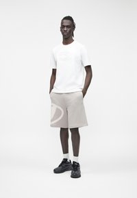 Mlad moški stoji z rokami v žepih, oblečen v belo majico s privzdignjenim logotipom, svetlo bež kratke hlače z velikim logotipom, bele nogavice in črne superge.
