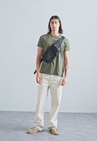 Must mustkott, mis on kantud üle keha, oliivroheline T-särk, hallid avarad püksid ja beežid sneakersid.